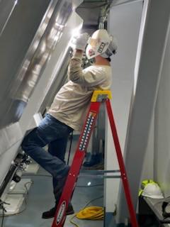 Technician inspecting an overhead HVAC or duct surface while positioned safely on an A-frame ladder.