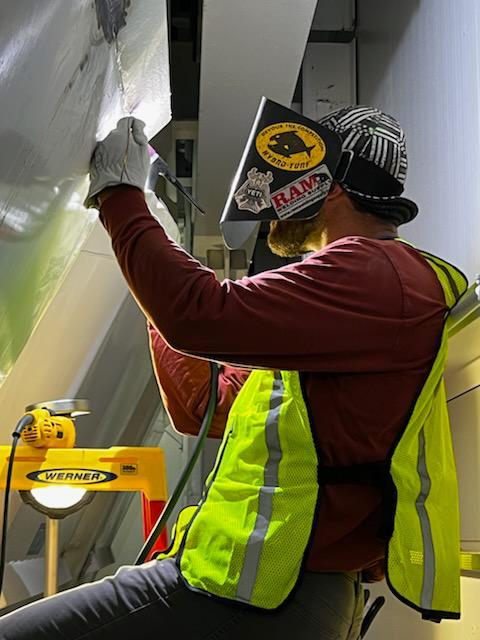 Technician performing close visual inspection on overhead ducting seam using hand tool and light.